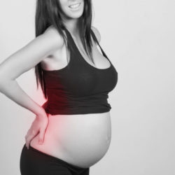 Brunette pregnant woman, with a strong pain, massaging her back ache. Indoors, over a grey wall.